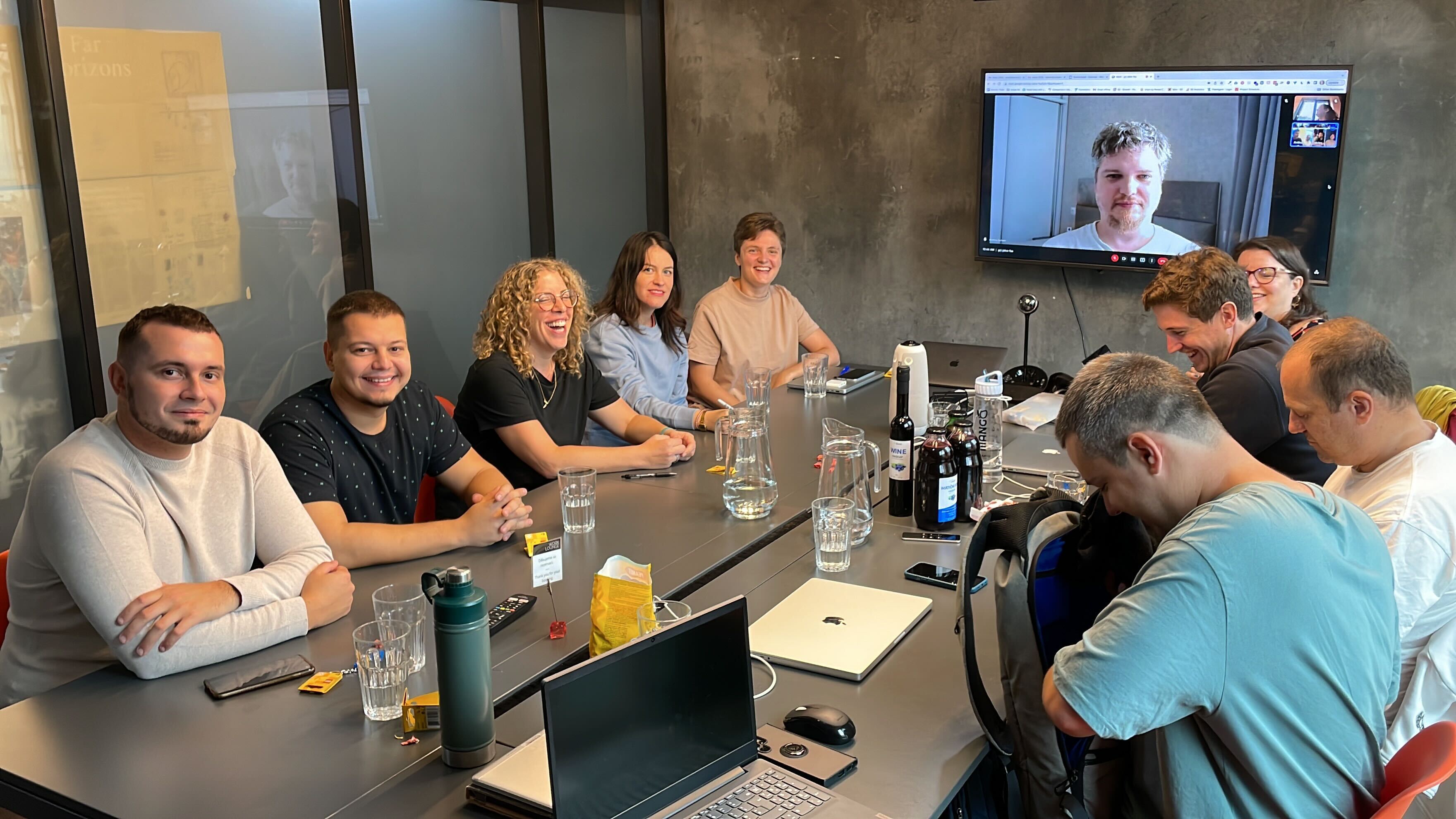 Andrey is on a big screen at the end of a table. The rest of the team are sitting round the table and smiling.